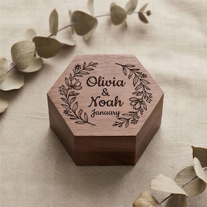 Könnte beinhalten: Eine sechseckige Holzbox mit floralem Kranzdesign und den Namen "Olivia & Noah" sowie dem Monat "Januar" auf dem Deckel eingraviert. Die Box hat eine warme braune Farbe und scheint aus Holz zu sein.