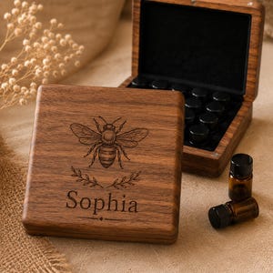 Può includere: Una scatola quadrata in legno con venature marrone scuro, caratterizzata da un'illustrazione di un'ape incisa sopra il nome Sophia. La scatola aperta rivela un interno nero con molteplici alloggiamenti circolari per flaconi in vetro ambrato.