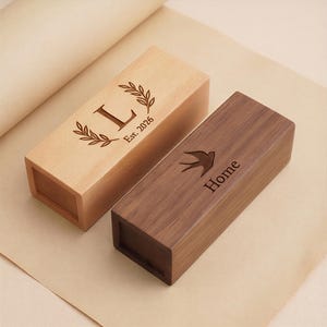 May include: Two wooden rectangular boxes. One is light-coloured with an engraved "L" inside a laurel wreath and the text "Est. 2026". The other is dark brown with a bird and the word "Home" engraved on the top.