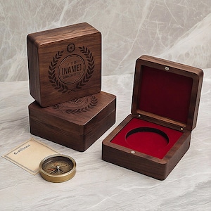 May include: Three wooden boxes with engraved text and a compass. The boxes are dark brown with a square shape. One box is open, revealing a red interior. A small, antique-style compass and a certificate are also present.