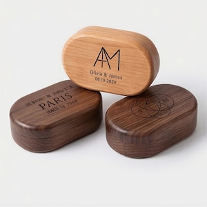May include: Three wooden, oval-shaped boxes. The top box is light brown with the initials "AM", names, and a date. The bottom two boxes are dark brown, one with the word "PARIS" and coordinates, and the other with interlocking circles and the letter "M".
