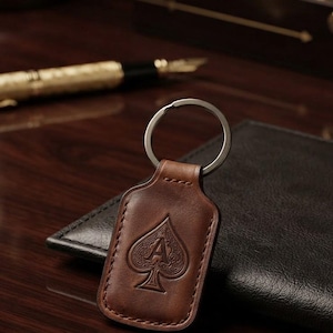 May include: A brown leather keyring with a rectangular shape and rounded corners, featuring a debossed spade symbol with the letter A in the centre. The leather is stitched along the edges and attached to a circular silver-toned metal split ring.