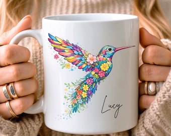 Op de afbeelding: Witte keramische mok met een kleurrijk kolibrie-ontwerp. De kolibrie is versierd met bloemen in verschillende kleuren. De naam "Lucy" staat in sierlijke letters op de mok.