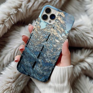 May include: A blue and white phone case featuring a winter landscape design. The case has a mosaic-like pattern with shades of blue, white, and gray, depicting trees and a sky. The case is held in a person's hand.