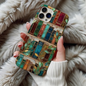 May include: A colorful phone case featuring a stained-glass design of bookshelves with books in shades of red, green, blue, and brown. The case has a gold outline and green leaf accents. The phone case is held by a person.