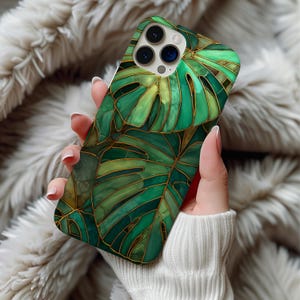 May include: A smartphone case featuring a stained-glass design of green monstera leaves with gold outlines. The case has a glossy finish and a protective design. The case is held in a person's hand.