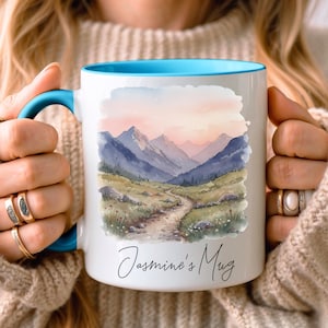 May include: White ceramic mug with a blue interior and handle. The mug features a watercolor painting of a mountain landscape with a path. The text "Jasmine's Mug" is written in cursive on the front.