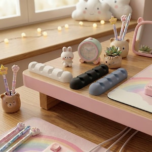May include: A desk setup with various accessories, including a white, black, and blue cable tidy, a small alarm clock, a bear-shaped pen holder, and a cat-shaped succulent pot. The desk has a pink surface and a wooden base.