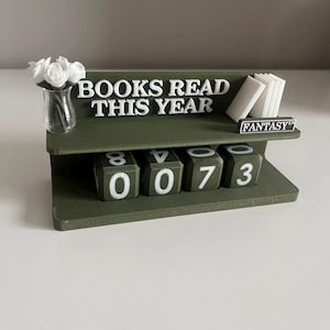 May include: A green bookshelf-style book counter with the words "BOOKS READ THIS YEAR" in white. It has a small vase with white flowers, a stack of white book replicas, and a counter showing "073". The word "FANTASY" is also visible.