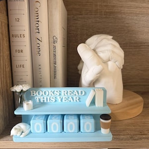 May include: A light blue plastic shelf-shaped calendar with the text BOOKS READ THIS YEAR. It features four rotating number blocks, a miniature white cat, a small vase with white flowers, a candle, and a coffee cup figurine.