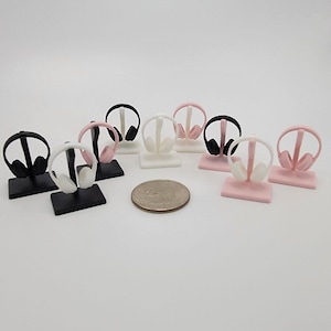 May include: Miniature headphone stands in black, white, and pink. Each stand holds a tiny headphone replica. The stands are rectangular and the headphones are in a variety of colors. A coin is next to the stands for size comparison.