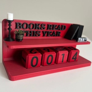 May include: A red shelf with the text "BOOKS READ THIS YEAR" and a set of rotating number blocks. The blocks display the number "2014". A small candle holder, a potted plant, and a stack of books are also on the shelf.