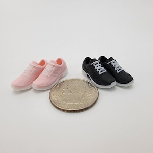 May include: Miniature pink and black sneakers with white laces are displayed next to a quarter. The pink sneakers are on the left, and the black sneakers are on the right. The sneakers are small, detailed, and appear to be made of plastic.