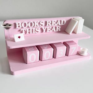 May include: A pink desk organizer with the text "BOOKS READ THIS YEAR". It features a shelf with miniature books, a martini glass, an envelope with a heart, and number blocks displaying "0037".