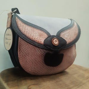 May include: A small, handcrafted shoulder bag with a unique design. The bag features a combination of materials, including a light gray top, pink snakeskin-textured panels, and black leather accents. A small tag hangs from the strap.