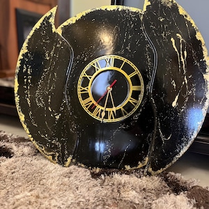 Può includere: Un orologio da parete decorativo nero e oro. Il quadrante presenta numeri romani e lancette dorate su sfondo nero. La cornice esterna ha una forma unica e astratta con accenti dorati.