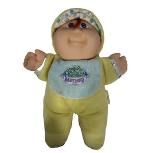 Peut inclure: Une poupée vintage Cabbage Patch Kids Babyland. La poupée a un corps en peluche jaune doux et porte une capuche et un bavoir assortis. Le bavoir porte le logo Babyland. La poupée a un visage en vinyle avec des yeux bleus et des cheveux roux.