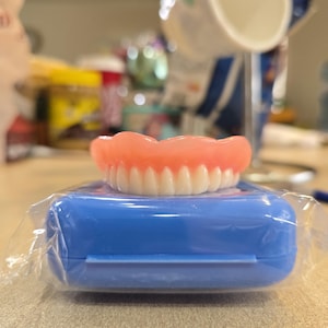 May include: A set of upper dentures with white teeth and a pink gumline, resting on a blue plastic case, wrapped in clear plastic. The dentures are the primary focus, suggesting a dental care or medical theme.