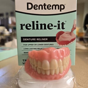 Puede incluir: Un juego de dentaduras postizas descansa sobre una almohadilla blanca frente a una caja de reliner de dentaduras Dentemp reline-it. La caja es de color verde claro y blanco con el nombre y la descripción del producto. Las dentaduras son rosas con dientes blancos.