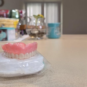 Puede incluir: Un juego de dentaduras postizas superiores con encías rosas y dientes blancos, apoyado sobre una almohadilla blanca dentro de un paquete de plástico transparente. Las dentaduras están enfocadas, el fondo borroso.
