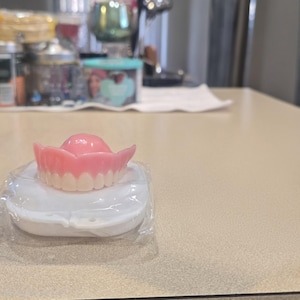 Puede incluir: Un juego de dentaduras postizas superiores con encías rosas y dientes blancos, colocado sobre una caja de plástico blanca. Las dentaduras están en un envoltorio de plástico transparente, sobre una mesa de color claro. El fondo está borroso.