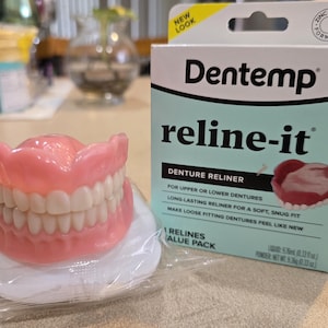 Può includere: Un set di dentiere con gengive rosa e denti bianchi, appoggiato su un tampone bianco. Una scatola di Dentemp reline-it per la riparazione delle dentiere è accanto alle dentiere. La scatola è azzurra e bianca con la scritta "reline-it".