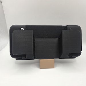 Pode incluir: Suporte de telefone de plástico preto com um padrão geométrico na parte de trás. O suporte tem duas presilhas laterais e um pequeno suporte retangular. O suporte é de cor bege claro. O suporte foi projetado para segurar um telefone.