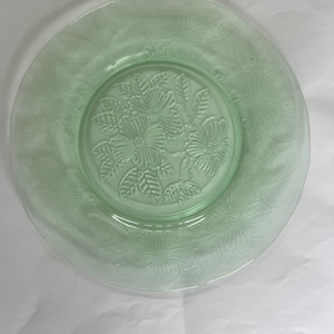 Puede incluir: Un plato de vidrio verde claro con un diseño floral. El plato presenta flores y hojas en relieve alrededor del borde y en el centro. El plato es redondo y tiene un borde ligeramente elevado.