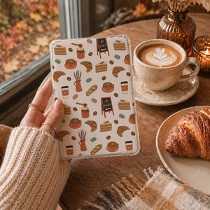 Könnte beinhalten: Transparente Handyhülle mit einem Café-Themenmuster mit Croissants, Kaffeetassen, Kuchenstücken und einem kleinen Café-Schild. Die Hülle wird in der Nähe eines Tisches mit einer Kaffeetasse, einem Croissant und einer Vase mit Trockenblumen gehalten.
