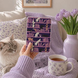 May include: A clear tablet case featuring a purple bookshelf design with various cats. The cats are in different poses and colors. The case is held in front of a gray cat, a purple blanket, and a floral teacup.