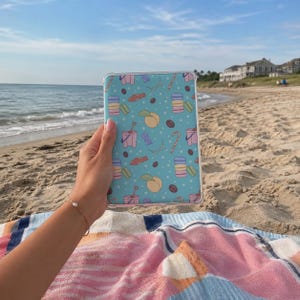 May include: A light blue tablet case with a repeating pattern of macarons, peaches, coffee beans, and candy canes. The case is held up against a beach scene with the ocean and sky in the background.