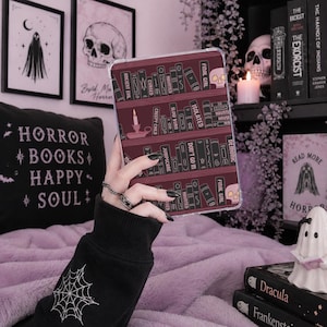 May include: A clear tablet case featuring a bookshelf design with horror-themed book titles and a skull. The case is held in front of a pillow with the words "HORROR BOOKS HAPPY SOUL" and framed artwork. A ghost figurine and books are also visible.