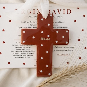 Peut inclure: Une croix en céramique de couleur terre cuite ornée de pois blancs et du prénom David inscrit en lettres dorées. La croix est suspendue par un ruban blanc et est placée sur une carte à pois rouges avec du texte en français.