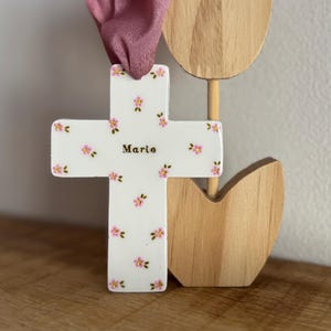 Puede incluir: Una cruz de cerámica blanca con un patrón repetitivo de pequeñas flores rosas con hojas verdes. El nombre Marie está impreso en letras doradas en el centro. Una cinta de color rosa empolvado está atada a través del agujero superior.