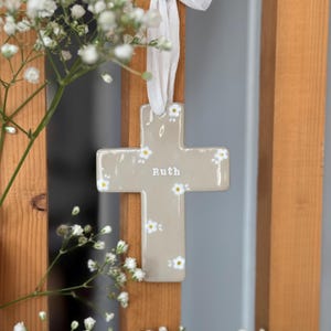 Peut inclure: Une croix en céramique vernissée de couleur taupe ornée d'un motif peint à la main de petites marguerites blanches aux cœurs jaunes. Le prénom Ruth est inscrit en lettres blanches au centre. Un ruban en tissu blanc passe par le haut.