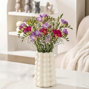 Puede incluir: Un jarrón de cerámica blanca con un diseño texturizado en forma de burbujas contiene un vibrante ramo de flores. El arreglo incluye flores moradas, rosas y blancas con tallos verdes. El jarrón está sobre una superficie blanca, con objetos decorativos en un estante al fondo.