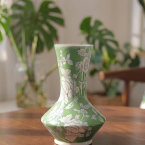 May include: A green ceramic vase with a white floral pattern. The vase has a flared base, a slender neck, and a slightly wider top. The vase is approximately 8 inches tall.