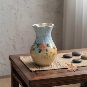 Pode incluir: Um vaso de cerâmica com fundo azul claro e borda ondulada. O vaso apresenta flores rosa pintadas à mão com folhas verdes e uma base amarela. O vaso é exibido em uma mesa de madeira.