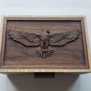 May include: Wooden box with a carved eagle design. The eagle is depicted with outstretched wings, centered on a dark brown wooden surface. The box has a light-colored wooden frame and a small, dark brown wooden pull.