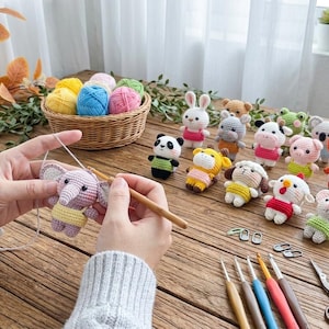 Könnte beinhalten: Eine Sammlung kleiner, bunter gehäkelter Tierfiguren, darunter ein Panda, ein Hase, ein Elefant und ein Koala. Ein Korb mit Garn, mehrere Metallhäkelnadeln und eine Schere sind zusammen mit den fertigen Spielzeugen auf einer Holzfläche angeordnet.