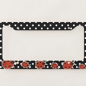 May include: Black license plate frame with white polka dots and red ladybug illustrations. The ladybugs have black spots and white antennae. The frame has a rectangular shape with rounded corners and holes for mounting.