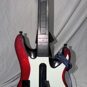 May include: A red glittery video game guitar controller featuring a white pickguard, a black neck with blue and orange fret buttons, and a coiled blue cable. The body includes a black strum bar and a button labeled Star Power at the base.