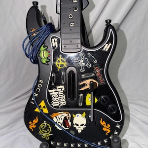May include: A black Guitar Hero video game controller shaped like an electric guitar, featuring a neck with the text ROCKS! and a body covered in various stickers including a skull, a tiger, and the Guitar Hero logo, with a blue coiled cable.