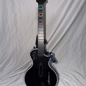 May include: A black Gibson branded video game guitar controller featuring five colored fret buttons in green, red, yellow, blue, and orange on the neck. The body includes a strum bar, a whammy bar, and circular control buttons on the front.