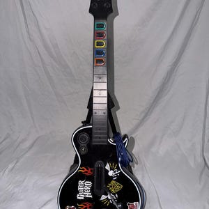 May include: A black Gibson Guitar Hero video game controller featuring five colored fret buttons on the neck, a strum bar, and various stickers on the body including flames, skulls, and the Guitar Hero logo. The controller is mounted on a stand.