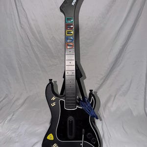 May include: A black Kramer Guitar Hero controller featuring five colored fret buttons on the neck, a whammy bar, a strum bar, and a central body with Guitar Hero branding, stickers, and a blue coiled cable attached to the guitar body.