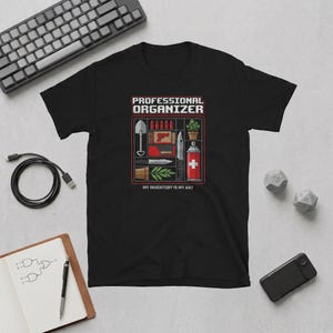 May include: A black t-shirt featuring a graphic print with the text "PROFESSIONAL ORGANIZER" and "MY INVENTORY IS MY ART". The design displays a grid of various items including a shovel, bullets, a potted plant, a knife, and a spray can.