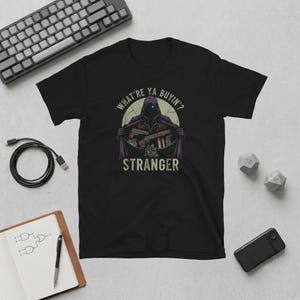 May include: A black short-sleeve t-shirt featuring a graphic of a hooded figure with a glowing blue eye opening a cloak to reveal various weapons. The text "WHAT'RE YA BUYIN'?" and "STRANGER" is printed in a light beige colour above and below the figure.