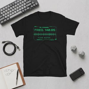 May include: A black short-sleeve t-shirt featuring a green graphic print of a digital frequency display. The text reads "FREQ. 140.85" above a sound wave pattern and the words "PLEASE RESPOND..." enclosed within a rectangular border.