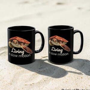 May include: Two black ceramic mugs featuring a realistic image of a sea turtle swimming. Below the turtle, the white text reads Living in slow motion. Each mug has a curved handle on the right side and a smooth, glossy finish on the exterior surface.